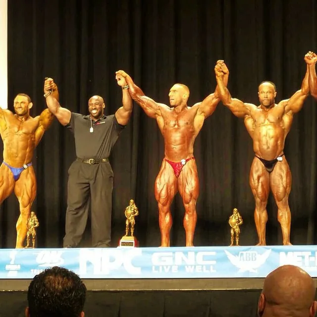 Dom Cardone having his arm raised by an official during the awards at an NPC show, three other competitors alongside him.