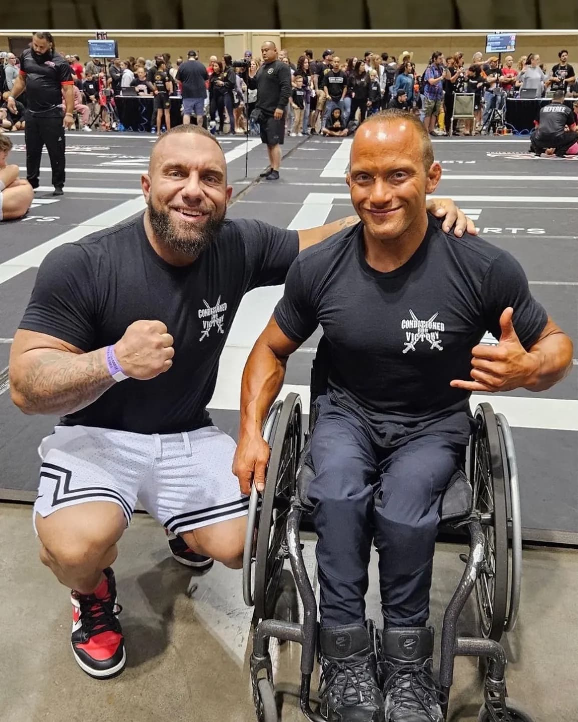 Dom Cardone crouching beside a coaching client in a wheelchair at a competition venue, both in team shirts.