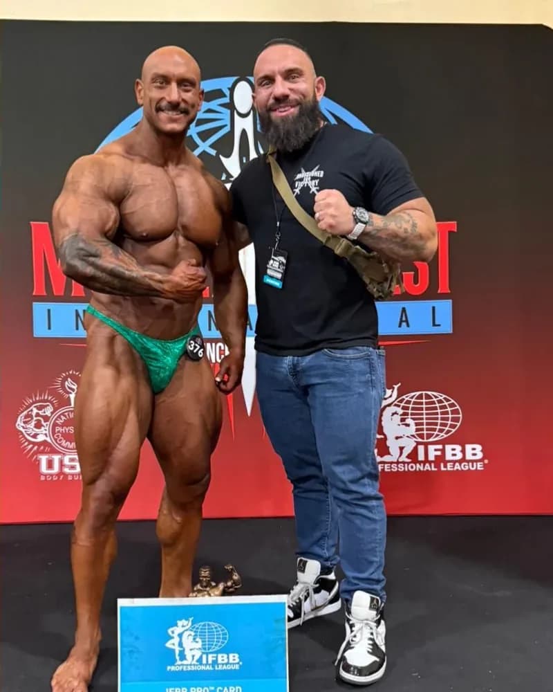 Dom Cardone on stage with a coaching client in contest condition, an IFBB pro card sign at their feet.